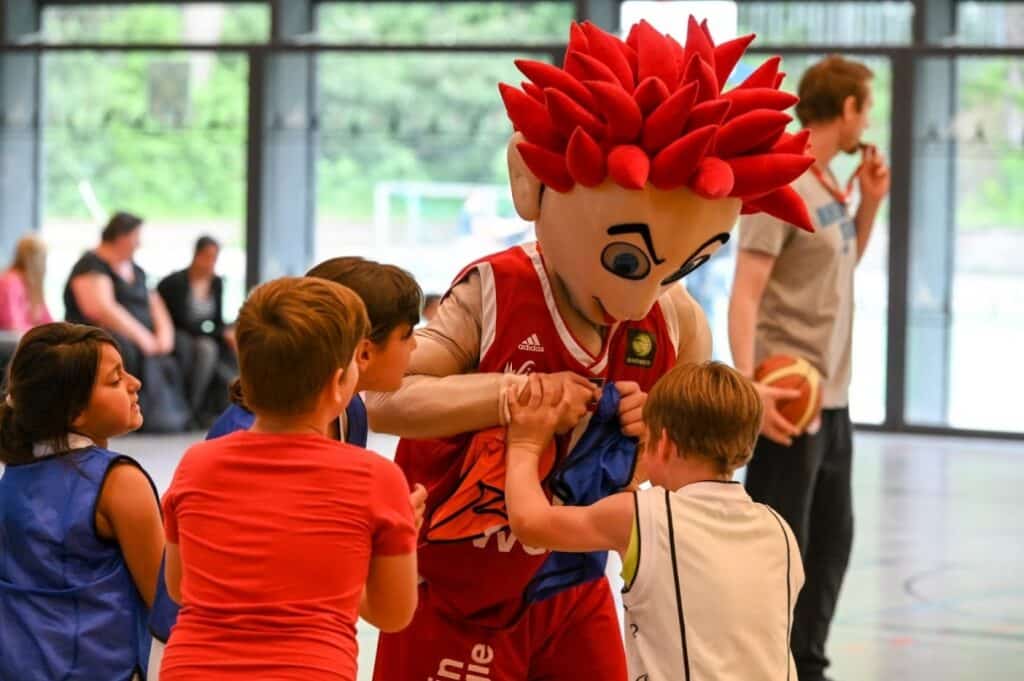 Maskottchen der RheinStars Köln mit roten Haaren umarmt Kinder in einer Sporthalle.