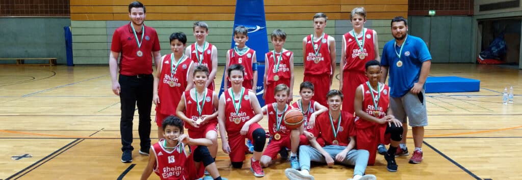 Kinderbasketballmannschaft RheinStars Köln mit Medaillen in Sporthalle posiert.