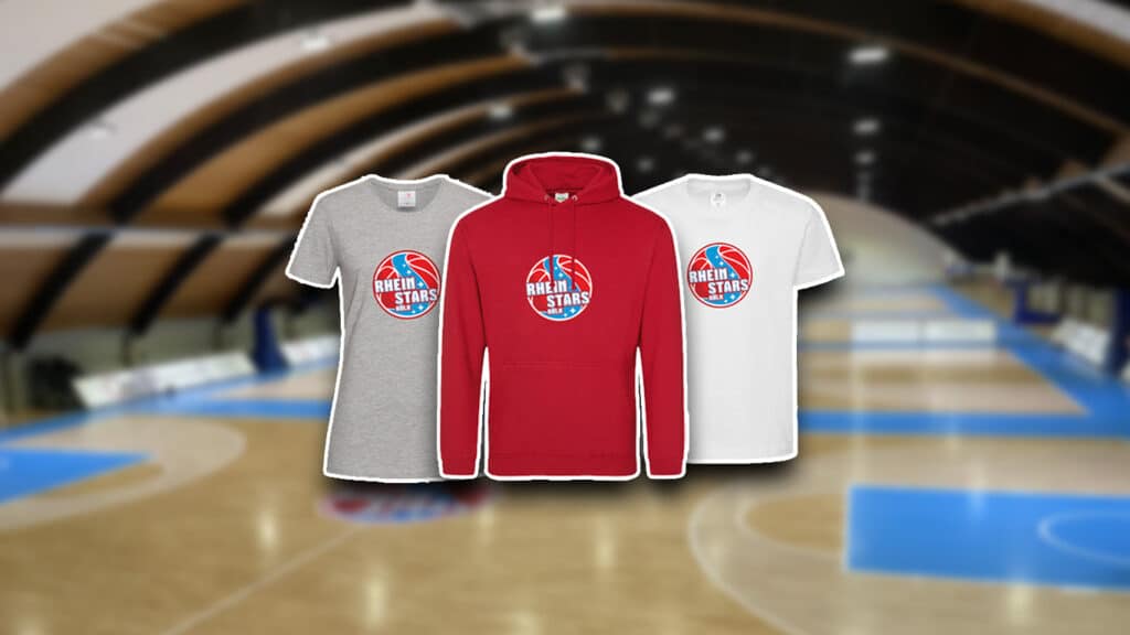 Drei RheinStars Köln Merchandise-Artikel auf Basketballplatz: graues T-Shirt, rotes Hoodie, weißes T-Shirt.