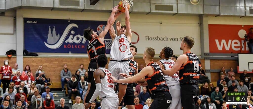 Basketballspiel der RheinStars Köln: Spieler in Weiß springt zum Wurf, schwarze Gegenspieler verteidigen.
