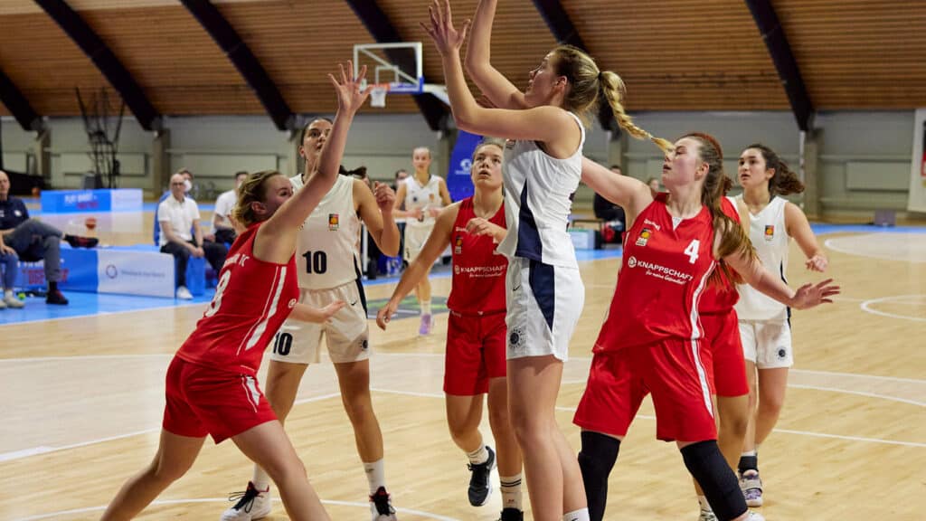 Basketballspiel der RheinStars Köln: Spielerin in Weiß wirft auf den Korb, während rote Gegnerinnen verteidigen.