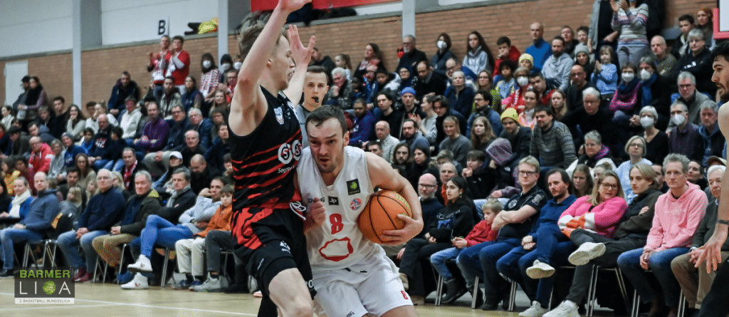 Basketballspiel der RheinStars Köln: Spieler in weißem Trikot dribbelt, Gegenspieler verteidigt, Zuschauer im Hintergrund.